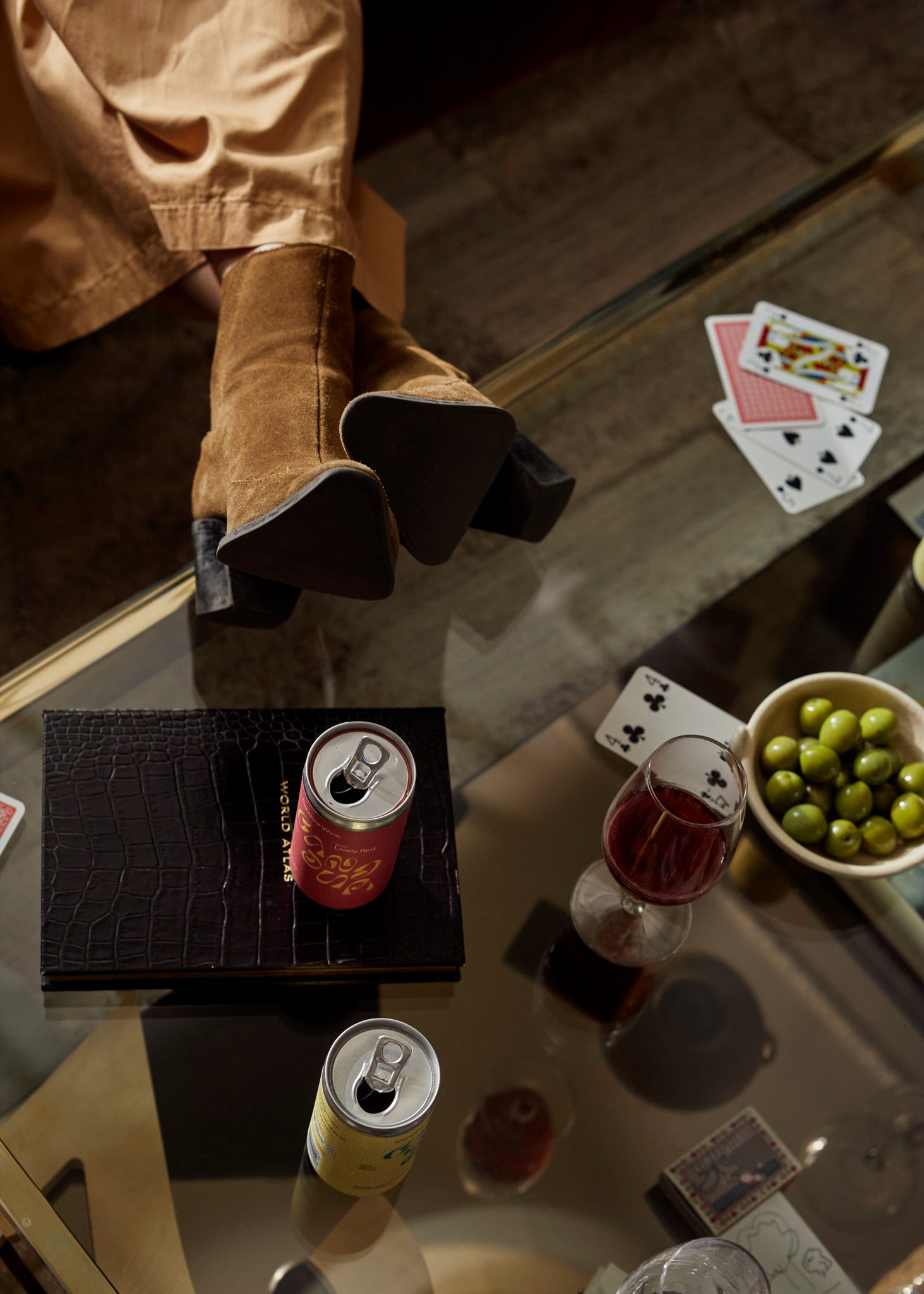 Woman wearing brown boots and resting her feet on a coffee table that holds a can of Candour red wine, a can of Candour white wine, cards, a book, and a bowl with green olives