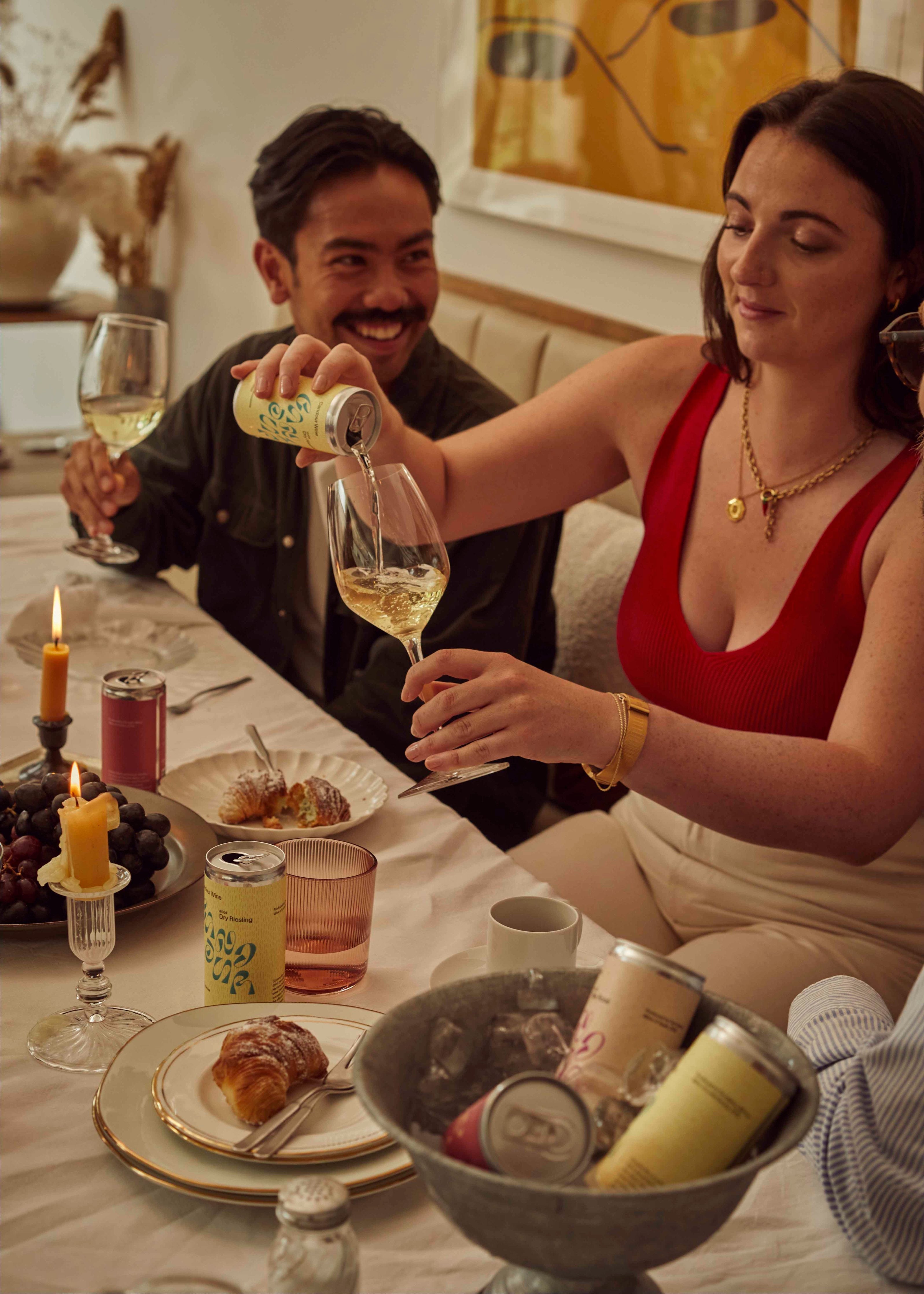 Woman seated at a table pouring white wine into a glass while her friend holds another glass and laughs. The remains of a nice dinner are on the table.
