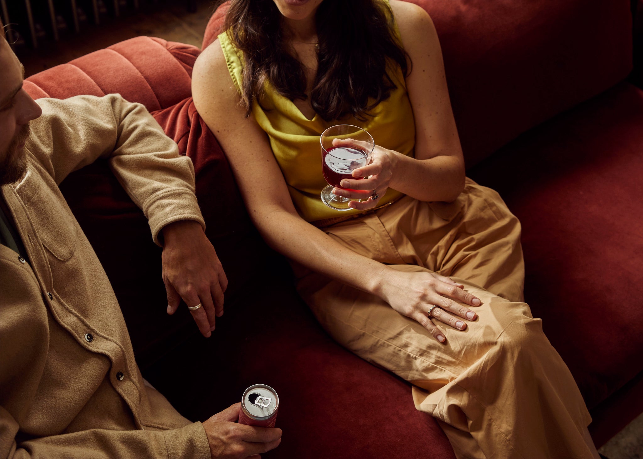 A woman and a man seating on a red velvet sofa. He has his elbow on the back of the sofa and is holding a can of Candour red wine. She has her hand over her crossed legs and is drinking a glass of red wine.