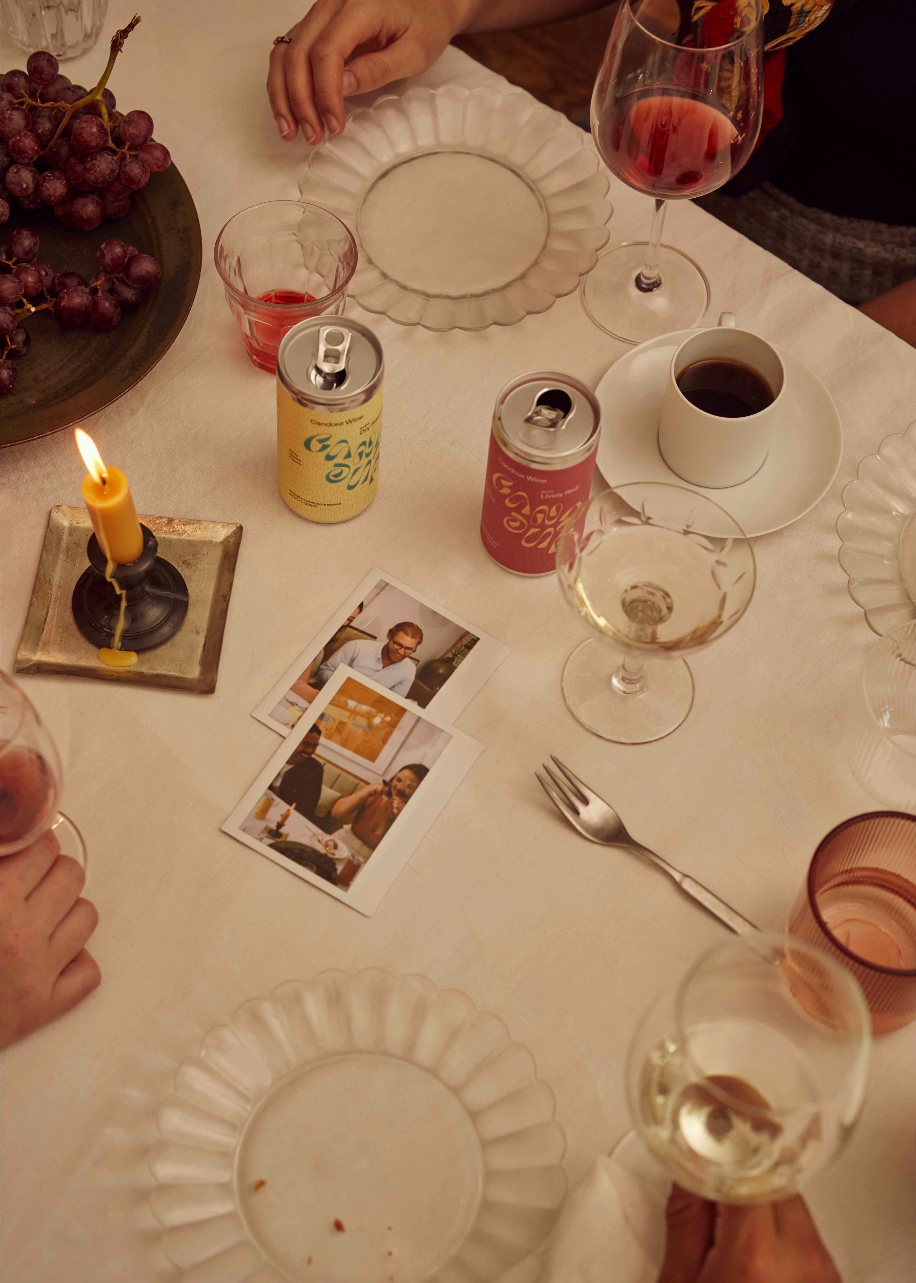 A can of red and a can of white wine on the table, with a poured glass of white wine and two polaroid pictures of friends.